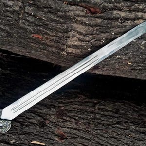 May include: A long, silver sword with a decorative handle and a purple grip. The blade has three parallel grooves running along its length. The sword is set against a dark, textured wooden background.