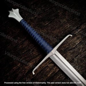 May include: A medieval sword with a silver blade and guard, and a blue leather-wrapped handle. The hilt is silver with a decorative pommel. The sword is set against a dark wood background. The text "Vikingscollectionit" is visible.