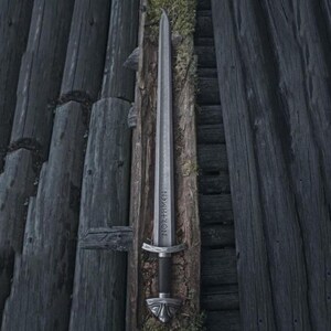 May include: A silver sword with the word "NORTHMEN" etched on the blade, resting against weathered wooden planks. The sword features a black handle and ornate silver hilt and pommel. The background is a dark, rustic wooden structure.