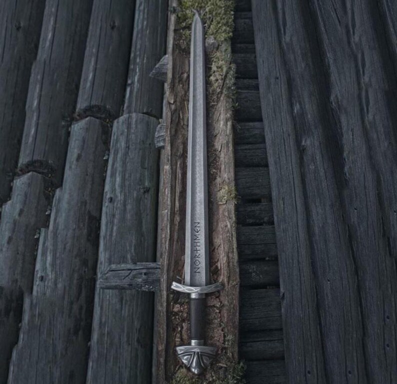 May include: A silver sword with the word "NORTHMEN" etched on the blade, resting against weathered wooden planks. The sword features a black handle and ornate silver hilt and pommel. The background is a dark, rustic wooden structure.
