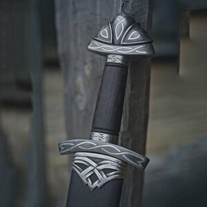 May include: A close-up of a sword with a dark wooden handle and silver accents. The hilt and pommel feature intricate designs. The blade is not visible. The sword is set against a weathered wooden background.
