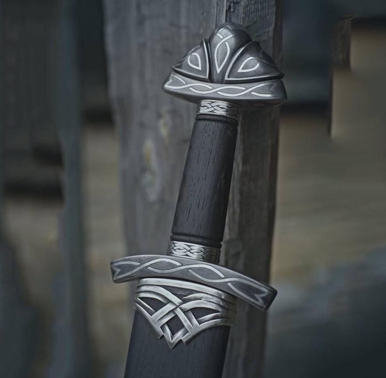 May include: A close-up of a sword with a dark wooden handle and silver accents. The hilt and pommel feature intricate designs. The blade is not visible. The sword is set against a weathered wooden background.