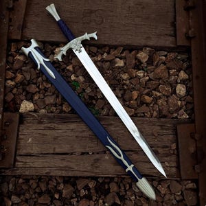 May include: A long sword with a double-edged steel blade, a silver-toned metal crossguard featuring a central emblem, and a dark blue grip. The matching dark blue scabbard has silver-toned metal fittings at the throat and the tip.