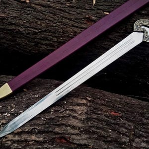 May include: A silver sword with a decorative handle and a purple sheath with a gold tip rests on a dark, textured surface. The sword's blade has a sharp point and several parallel grooves. The handle features an ornate design.
