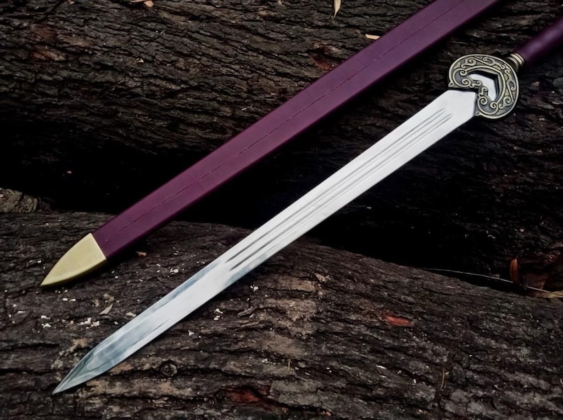 May include: A silver sword with a decorative handle and a purple sheath with a gold tip rests on a dark, textured surface. The sword's blade has a sharp point and several parallel grooves. The handle features an ornate design.