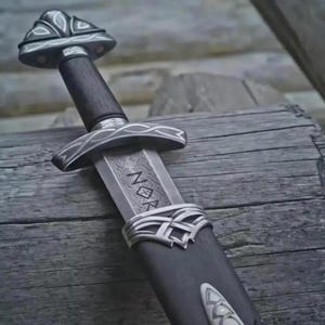 May include: A detailed image of a Viking sword with a dark wooden handle and a silver-colored guard. The blade has etched markings. The sword rests on a weathered wooden surface, showcasing its intricate design and craftsmanship.