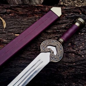 May include: A decorative sword with a polished steel blade and a detailed bronze hilt. The handle and scabbard are a deep purple color. The sword is resting on a dark brown wooden surface.