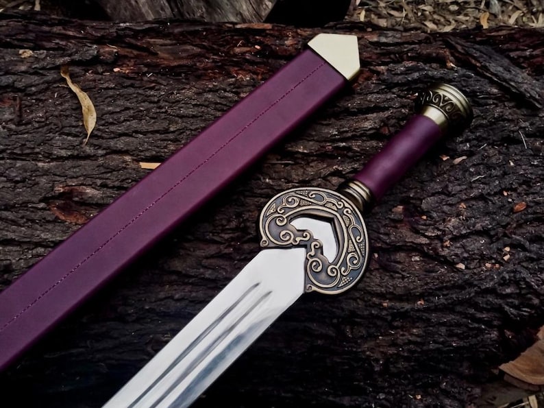 May include: A decorative sword with a polished steel blade and a detailed bronze hilt. The handle and scabbard are a deep purple color. The sword is resting on a dark brown wooden surface.