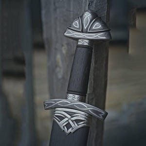 May include: A close-up of a sword with a dark wooden handle and silver accents. The hilt and guard feature intricate Celtic knot designs. The blade is sheathed in a dark scabbard, and the sword is leaning against a wooden surface.