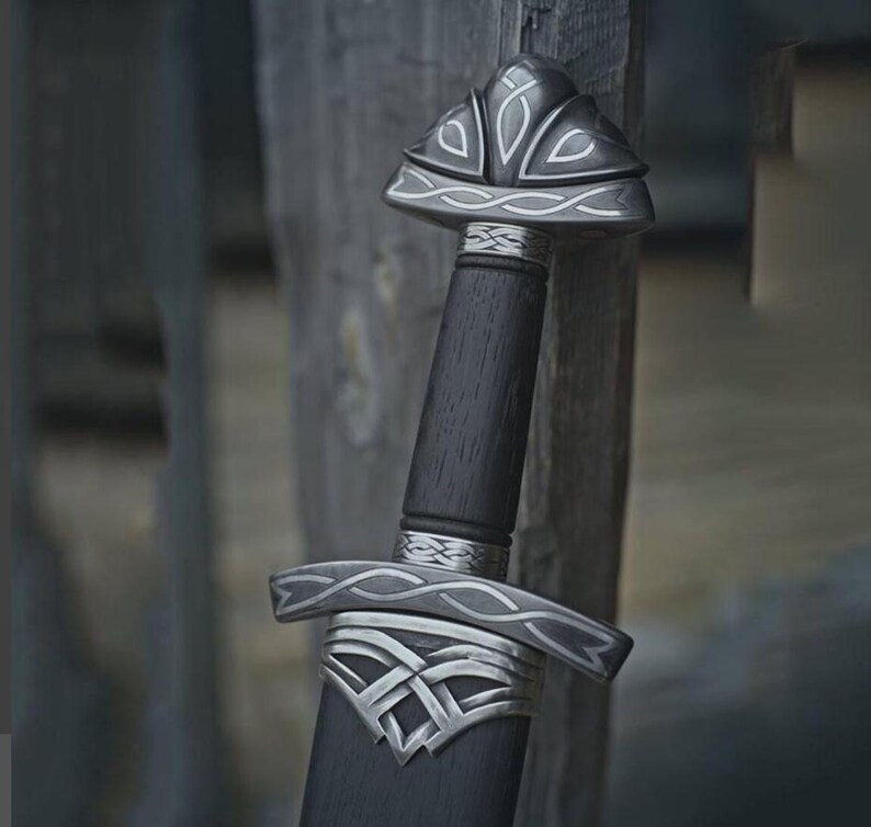 May include: A close-up of a sword with a dark wooden handle and silver accents. The hilt and guard feature intricate Celtic knot designs. The blade is sheathed in a dark scabbard, and the sword is leaning against a wooden surface.