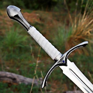Puede incluir: Una espada de estilo medieval con una hoja y guarda plateadas. El mango está envuelto en material blanco. Una pequeña gema azul está engastada en la guarda. La espada tiene una punta y está sobre un fondo verde borroso.