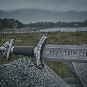 May include: A detailed image of a sword with the word "NORTHMEN" etched on the blade. The sword features a dark wooden handle and ornate silver-colored accents. The background shows a natural outdoor setting with a lake and overcast sky.