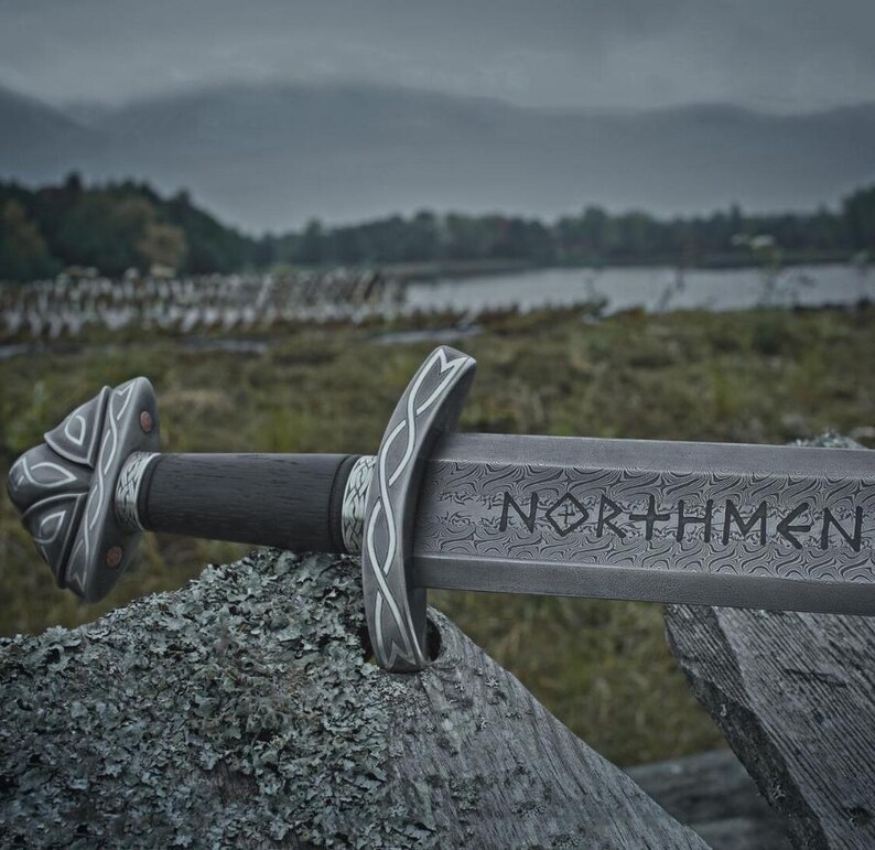 May include: A detailed image of a sword with the word "NORTHMEN" etched on the blade. The sword features a dark wooden handle and ornate silver-colored accents. The background shows a natural outdoor setting with a lake and overcast sky.