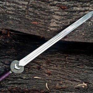 May include: A silver sword with a decorative heart-shaped hilt and a purple handle. The blade has three grooves running along its length. The sword is resting on a textured, dark brown wooden surface.