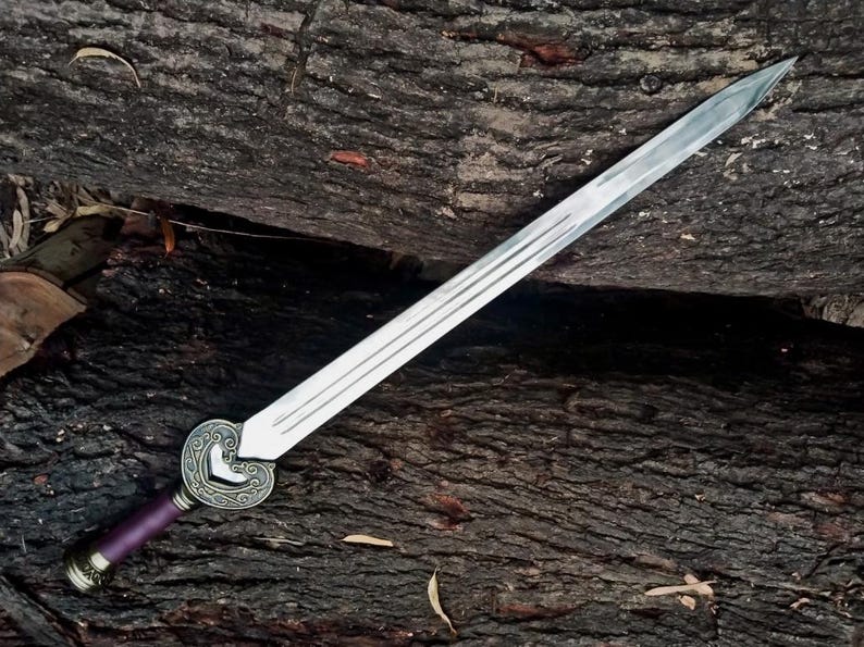 May include: A silver sword with a decorative heart-shaped hilt and a purple handle. The blade has three grooves running along its length. The sword is resting on a textured, dark brown wooden surface.