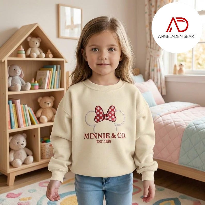 A young girl wearing a white sweatshirt with a Minnie Mouse design stands in a bedroom with a bookshelf and stuffed animals in the background.