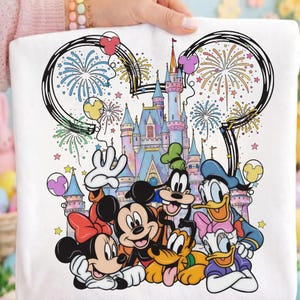 Puede incluir: Camiseta blanca con personajes de Disney: Mickey Mouse, Minnie Mouse, Donald Duck, Daisy Duck, Goofy y Pluto frente a un castillo colorido. El diseño incluye fuegos artificiales, globos y las orejas de Mickey Mouse.