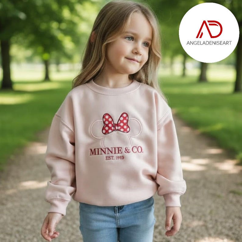 A young girl wearing a pink sweatshirt with a Minnie Mouse logo and the text "MINNIE & CO EST. 1979" is walking on a path in a park.
