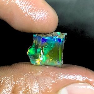May include: A vibrant, raw opal gemstone with a mix of colors, including green, blue, and yellow. The rough stone is held between fingers, showcasing its natural, unpolished state. The opal's surface reflects light, creating a shimmering effect.
