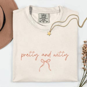 May include: Cream-colored t-shirt with the words "pretty and witty" in a cursive font, along with a bow graphic. A gold star pendant necklace and a brown hat are also visible. The shirt is a Comfort Colors brand, size XL.