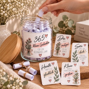 Puede incluir: Un frasco de vidrio transparente con tapa de madera lleno de pequeños rollos de papel blanco atados con cordel. La etiqueta dice 365 kleine Gedanken, junto a varias tarjetas a juego con ilustraciones florales y texto en alemán.