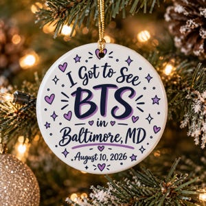 May include: A round white ceramic ornament featuring the text I Got to See BTS in Baltimore, MD and August 10, 2026 in dark blue and purple lettering. The design is decorated with small purple hearts and stars, and includes a gold hanging string.