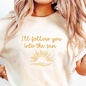 May include: A cream-colored short-sleeve t-shirt featuring the yellow text "I'll follow you into the sun" printed on the chest, accompanied by a graphic of a rising sun over wavy lines, all rendered in a matching yellow tone.