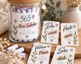 May include: A clear glass jar with a bamboo lid filled with small rolled white paper notes. The jar label reads 365 kleine Gedanken für ein besonderes Jahr. Four rectangular cards with floral illustrations and German text are displayed nearby.