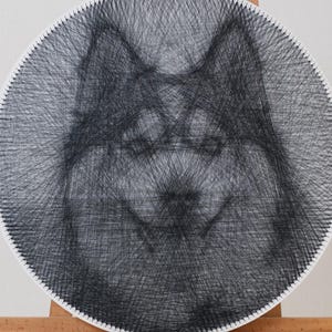 Può includere: Un'opera d'arte circolare con filo raffigurante il volto di un cane, creata con filo nero su sfondo bianco. L'opera è esposta su un cavalletto di legno, evidenziando i dettagli del volto del cane.
