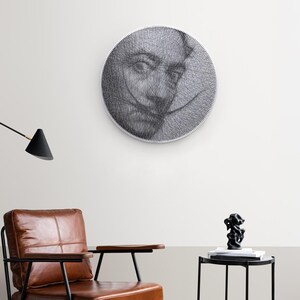 Könnte beinhalten: Runde Wandkunst mit einem Porträt, das mit schwarzem Faden auf weißem Hintergrund erstellt wurde. Das Kunstwerk hängt über einem braunen Ledersessel mit schwarzem Metallrahmen. Ein schwarzer Beistelltisch mit einer Skulptur befindet sich neben dem Stuhl.