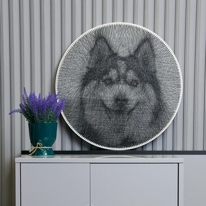 May include: Circular string art depicting a dog's face in black thread, set against a white backdrop. The artwork hangs on a grey wall. A teal vase with purple flowers is on a grey cabinet below.