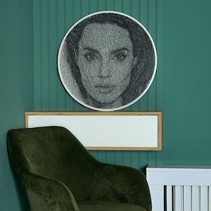 May include: A round, black and white string art portrait of a woman's face hangs on a teal wall. The artwork is framed in a white circle. Below the artwork is a white rectangular frame with a wooden border. A green velvet chair is in the foreground.