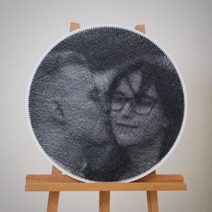 Puede incluir: Un retrato redondo de arte de hilo de dos personas, creado con hilo negro sobre una base circular con borde blanco. La obra de arte se exhibe en un caballete de madera, mostrando el detallado trabajo de hilo que forma los rostros.