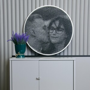 Pode incluir: Um retrato redondo de arte de fio de duas pessoas, com uma borda branca, está pendurado em uma parede cinza listrada. Um vaso verde-azulado com flores roxas está em um armário cinza. O armário tem duas portas.