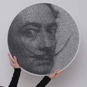 Pode incluir: Um retrato redondo de arte de fio de um homem com um bigode distinto, criado com fio preto em uma moldura circular branca. A obra de arte é segurada por duas mãos, mostrando o design e os detalhes intrincados.