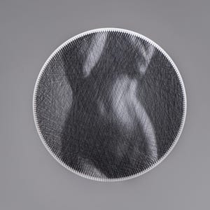 Pode incluir: Uma obra de arte circular em fio preto e branco, retratando uma figura humana. A obra de arte é feita com fio preto sobre uma base circular branca. O fundo é cinza.