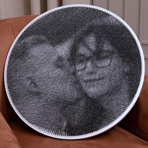 Puede incluir: Un retrato redondo de arte de hilo en blanco y negro. La obra de arte presenta dos rostros, creados con hilo negro sobre un marco circular blanco. La pieza está montada sobre una superficie marrón, mostrando el intrincado diseño de hilo.