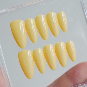 May include: A set of ten yellow press-on nails in a clear plastic case. The nails are oval-shaped and have a glossy finish. The color is a soft, pastel yellow.