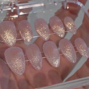 May include: A set of ten artificial nails in a clear plastic case. The nails are oval-shaped and have a light pink base with a glitter gradient from the tips. The glitter is a mix of gold and silver, creating a shimmering effect.