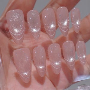 Puede incluir: Un juego de diez uñas artificiales en un tono rosa claro. Cada uña presenta un acento brillante y un delicado diseño de líneas blancas cerca de la punta. Las uñas son de forma ovalada y están listas para usar.