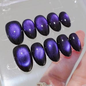May include: A set of ten press-on nails featuring a deep purple cat-eye magnetic polish effect. Each nail has an oval shape with a dark base color and a shimmering, bright violet center that creates a reflective, dimensional appearance on the surface.