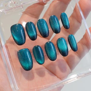 Könnte beinhalten: Ein Set aus zehn oval geformten, teal-farbenen Kunstnägeln. Die Nägel haben ein glänzendes Finish mit einem schimmernden Cat-Eye-Effekt. Sie sind in einer transparenten Kunststoffbox angeordnet und sofort einsatzbereit.