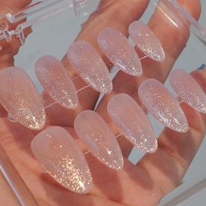 May include: A set of ten artificial nails in a clear plastic case. The nails are oval-shaped with a light pink base and a glitter gradient from the tips. The glitter transitions from a gold to a silver hue.
