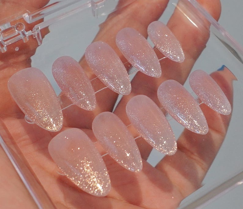 May include: A set of ten artificial nails in a clear plastic case. The nails are oval-shaped with a light pink base and a glitter gradient from the tips. The glitter transitions from a gold to a silver hue.