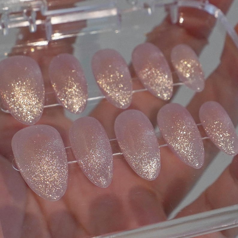 May include: A set of ten artificial nails in a clear plastic case. The nails are oval-shaped with a light pink base and a glittery gold gradient effect. The nails are designed for cosmetic use.