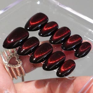 May include: A set of ten almond-shaped press-on nails featuring a deep black base color with a shimmering, magnetic red cat-eye effect running diagonally across the center of each nail, creating a glossy, reflective finish on the surface.