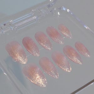 May include: A set of ten almond-shaped, press-on nails. The nails are a soft pink color with a glitter gradient, transitioning from a dense shimmer at the tips to a translucent base. They are displayed in a clear plastic case.