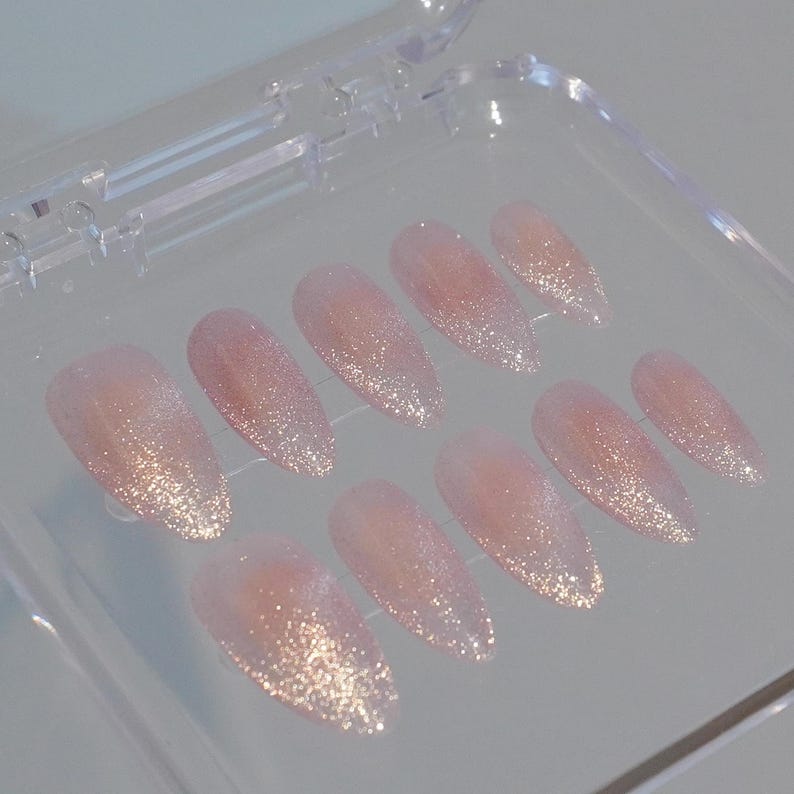 May include: A set of ten almond-shaped, press-on nails. The nails are a soft pink color with a glitter gradient, transitioning from a dense shimmer at the tips to a translucent base. They are displayed in a clear plastic case.