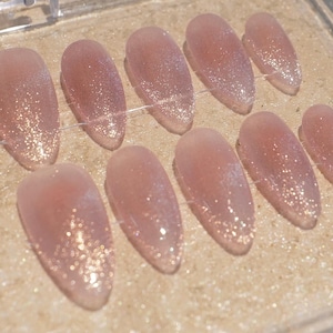 May include: A set of oval-shaped, light pink press-on nails with a glitter gradient. The nails are arranged in a row, showcasing a shimmering effect. The glitter transitions from a concentrated base to a subtle tip.