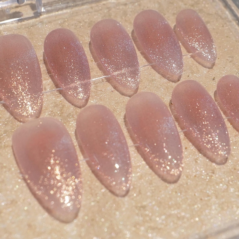 May include: A set of oval-shaped, light pink press-on nails with a glitter gradient. The nails are arranged in a row, showcasing a shimmering effect. The glitter transitions from a concentrated base to a subtle tip.